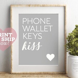 May include: Framed print with a gray background and white text that reads "PHONE WALLET KEYS kiss" with a white heart. A pink heart with the words "PRINT OR SHIP now!" is in the corner. A small plant in a white pot is on the right.