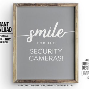 May include: A grey printable sign with white text that reads "smile for the security cameras!"  The sign is framed in a light brown wood frame. The text "*TRULY* ORIGINAL DESIGNS! {BE CREATIVE. DON'T COPY.} BATSHIT Craftie" is also included in the image.