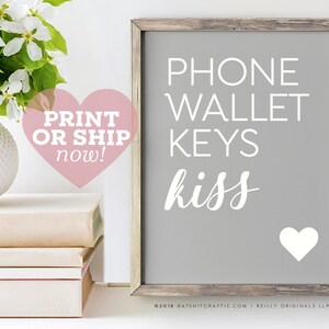 May include: A gray framed sign with white text that reads "PHONE WALLET KEYS kiss" with a white heart at the bottom. The sign is on a white surface with a stack of books and a white vase with white flowers.