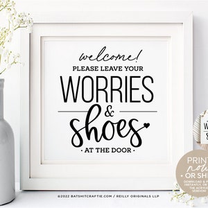 Puede incluir: Un cartel blanco con texto negro que dice "welcome! Please leave your worries & shoes at the door." El cartel está enmarcado en un marco blanco y está sobre una superficie blanca. Hay un pequeño jarrón blanco con flores blancas en el fondo.
