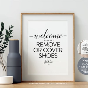 Puede incluir: Un cartel imprimible en blanco y negro que dice "Bienvenido, por favor, quítate o cubre los zapatos". El cartel está enmarcado en un marco de madera claro y está sobre una mesa con un jarrón de vegetación.