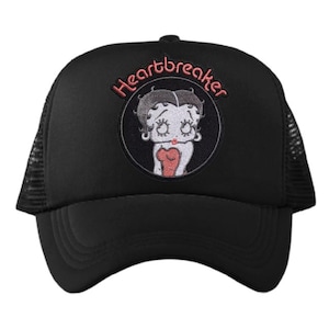 Puede incluir: Gorra de camionero negra con un parche circular con una imagen de dibujos animados de una mujer y la palabra "Heartbreaker" en rosa. La gorra tiene una visera curva y una parte trasera de malla.