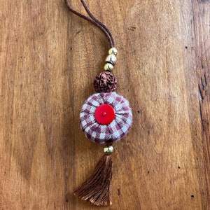 Peut inclure: Un porte-clés en tissu à carreaux brun et blanc avec un bouton rouge au centre. Le porte-clés est doté d'un cordon brun et de perles dorées.
