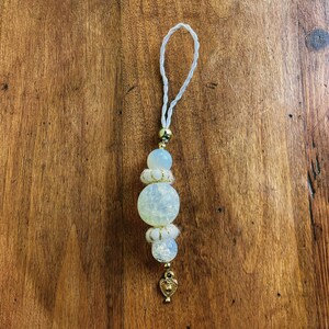 Peut inclure: Un charme en perles blanches et dorées avec un pendentif en forme de cœur. Le charme est attaché à une ficelle blanche.