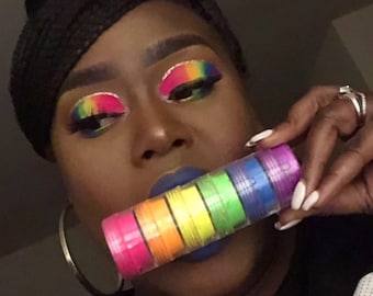 May include: A stack of rainbow-colored cosmetic pigments in small, clear containers. The makeup is held in front of a person with rainbow eye makeup and blue lipstick. The containers are arranged in a rainbow gradient, from pink to purple.