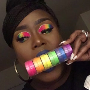 May include: A stack of rainbow-colored cosmetic pigments in small, clear containers. The makeup is held in front of a person with rainbow eye makeup and blue lipstick. The containers are arranged in a rainbow gradient, from pink to purple.