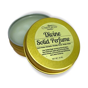 Puede incluir: Una lata de metal dorada con tapa que contiene un perfume sólido. La etiqueta dice "Divine Solid Perfume" con las notas de olor "Romántico, Amaderado, Floral, Ámbar, Almizcle". La etiqueta también incluye instrucciones de uso y el peso neto de 30 ml.