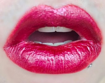 Pode incluir: Close-up de lábios abertos com batom vermelho brilhante. Os lábios são brilhantes e os dentes são brancos. Um pequeno piercing labial prateado é visível no lábio inferior.