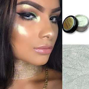Puede incluir: Una mujer con un maquillaje de ojos llamativo, que incluye sombra de ojos verde y delineador negro, e iluminador brillante. Se ve un frasco de "Diamond Highlighter", junto con un primer plano de la textura del producto. Un collar de gargantilla dorado.