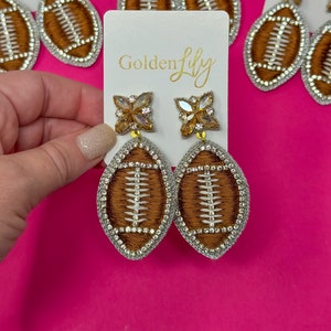 May include: A pair of brown and white football-shaped earrings with rhinestones. The earrings are hanging from a white card with the text "Golden Lily" printed on it.