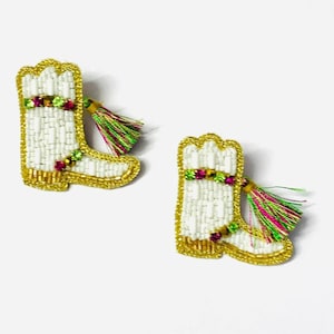 May include: A pair of white beaded cowboy boot earrings with gold trim and colourful gemstone accents. Each boot has a small tassel with pink, green, and yellow threads. The earrings are set against a white background.