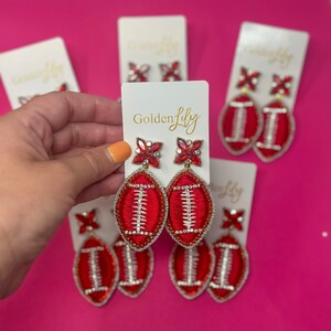 May include: Red and white football-shaped earrings with a beaded border and crystal accents. The earrings are displayed on white cards with the "Golden Lily" logo. The background is a bright pink.