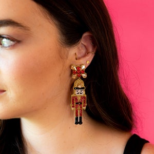 May include: A pair of festive, beaded earrings featuring a nutcracker design. The earrings have a red, gold, and black color scheme, with a flower-shaped top. The nutcracker figure is detailed with a hat and uniform.