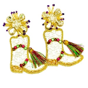 Puede incluir: Un par de pendientes de bota blancos con cuentas, detalles dorados y borlas de colores. Los pendientes tienen un diseño de flor de cristal en la parte superior.