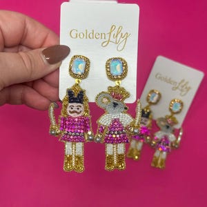 Puede incluir: Pendientes colgantes con diseño de cascanueces y rey ratón. Los pendientes están adornados con cuentas rosas, doradas y blancas, con una piedra cuadrada iridiscente en la parte superior. Fondo rosa.