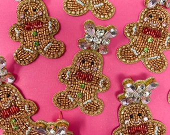 Op de afbeelding: Oorbellen in de vorm van peperkoekenmannetjes met een kralenontwerp. De oorbellen zijn bruin met rode strikjes en groene accenten. Elke oorbel heeft een kristallen versiering. De achtergrond is felroze.