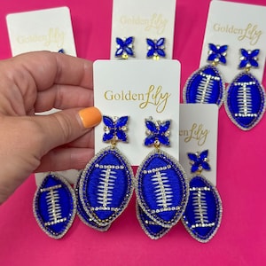 May include: Blue and white football-shaped earrings with rhinestones and a gold-coloured backing. The earrings are displayed on a white card with the text "Golden Lily" printed in gold.