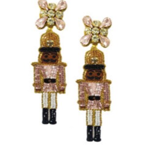 May include: Pink and gold beaded nutcracker earrings with crystal accents. The nutcracker is wearing a pink and gold uniform with a black hat and black boots.