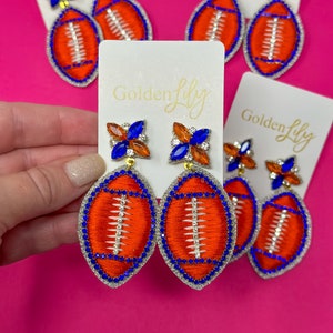 May include: A pair of football-shaped earrings with blue and orange rhinestones. The earrings are on a white card with the text "Golden Lily" printed on it.