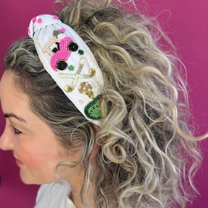 Op de afbeelding: Een wit hoofdband met een geborduurd ontwerp in roze, groen en goud met een golfkar en golfclubs.