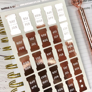 May include: A sheet of rose gold foil monthly calendar stickers with white text. The stickers are in the shape of a cylinder with a cut-out on the side. The months are January, February, March, April, May, June, July, August, September, October, November, and December.