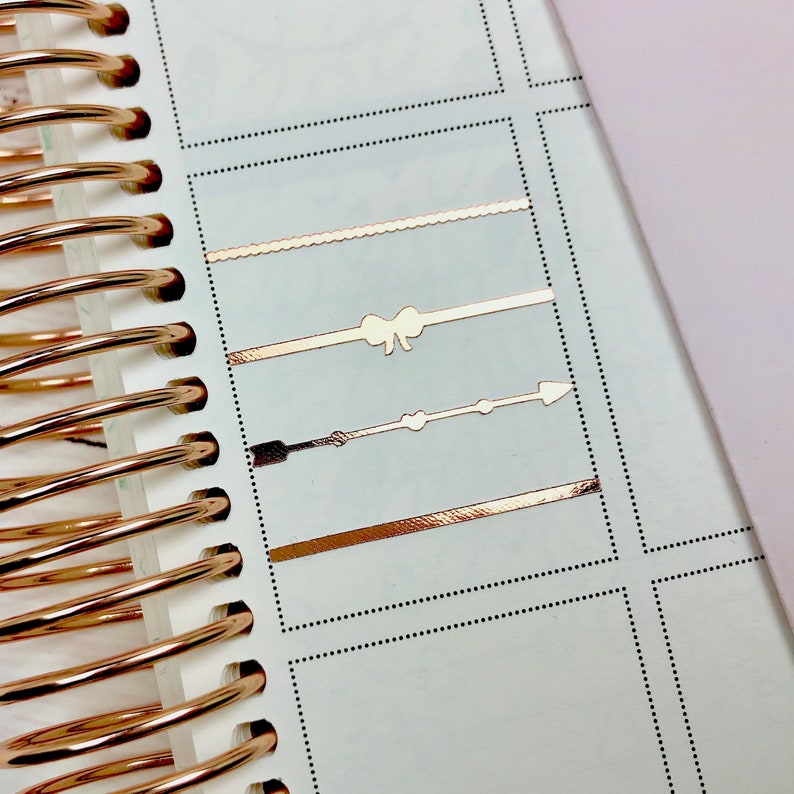 Op de afbeelding: Een close-up van een planner met vier gouden folie stickers. De stickers zijn een golvende lijn, een strik, een pijl en een rechte lijn.