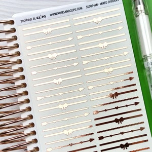 Op de afbeelding: Een vel rose gouden planner stickers met verschillende scheidingslijnontwerpen. De ontwerpen omvatten strikken, pijlen, lijnen en stippen. De stickers bevinden zich op een witte achtergrond en de tekst "notes & clips" en "www.notesandclips.com" staat bovenaan het vel gedrukt. De productcode "SS0094MI - MIXED DIVIDERS" staat rechtsboven gedrukt.