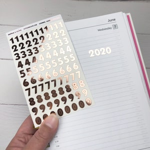 Op de afbeelding: Een vel met gouden cijfer stickers met de cijfers 0 tot en met 9. De cijfers zijn gerangschikt in rijen, waarbij elke rij hetzelfde cijfer bevat. Het vel wordt in een hand tegen een witte achtergrond gehouden.