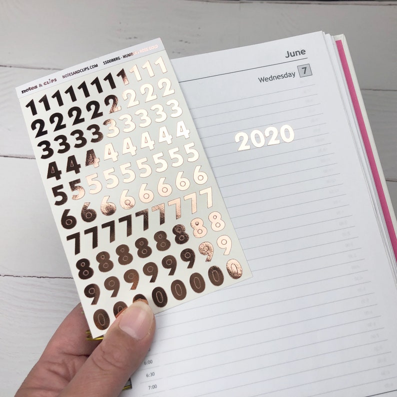 Op de afbeelding: Een vel met gouden cijfer stickers met de cijfers 0 tot en met 9. De cijfers zijn gerangschikt in rijen, waarbij elke rij hetzelfde cijfer bevat. Het vel wordt in een hand tegen een witte achtergrond gehouden.