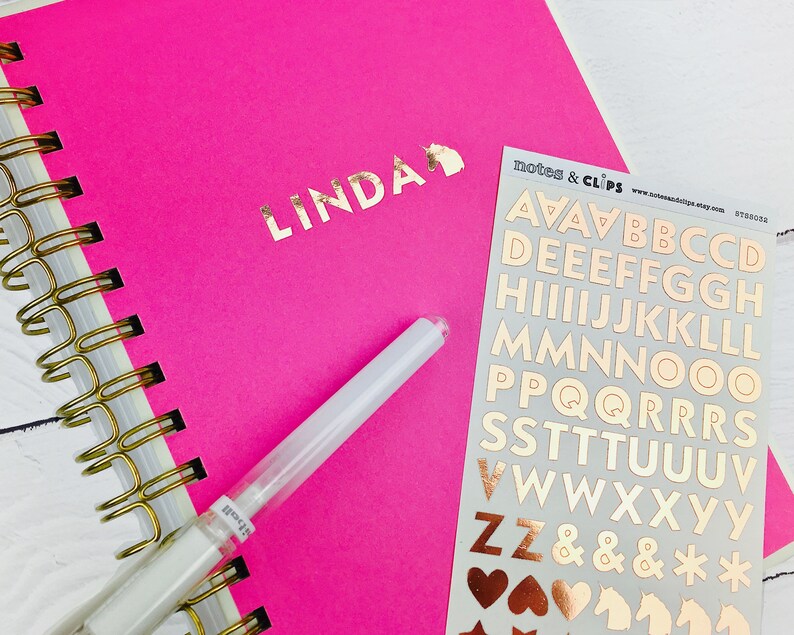 Op de afbeelding: Een roze notitieboek met een gouden spiraalbinding en de naam "LINDA" geschreven in gouden letters. Naast het notitieboek ligt een vel met gouden letter stickers. De stickers bevatten de letters A tot en met Z, ampersands, sterretjes en harten.