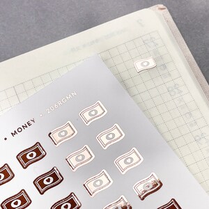 May include: A sheet of stickers with a rose gold finish. The stickers are shaped like money bills with a heart in the center. The text "MONEY. 206RGMM" is printed at the top of the sheet.