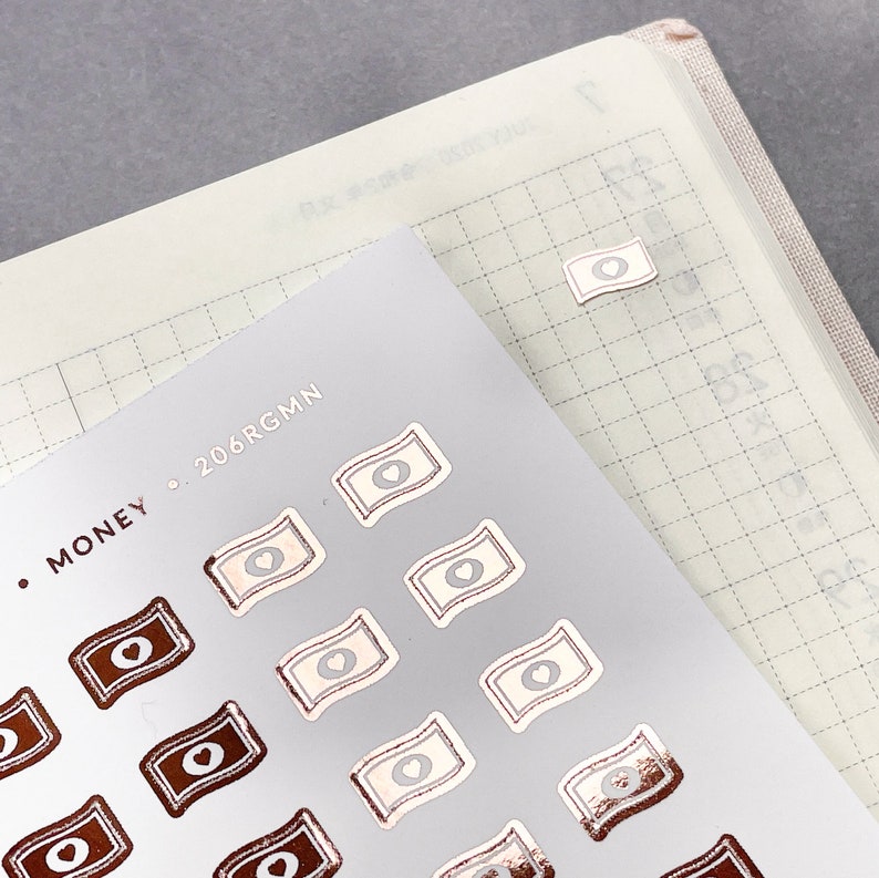 May include: A sheet of stickers with a rose gold finish. The stickers are shaped like money bills with a heart in the center. The text "MONEY. 206RGMM" is printed at the top of the sheet.