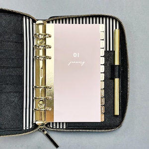 May include: A black zippered planner with a gold six-ring binder mechanism and a gold pen. The interior features black and white striped lining. A pale pink monthly divider page is inserted, labeled with the text 01 january and month tabs.