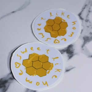 May include: Two round stickers with a white background and a yellow honeycomb design. The text "sweet like home" is written in yellow around the honeycomb. The stickers are on a white and gray marble surface.