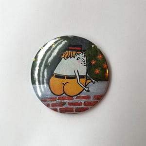 May include: A round button with a cartoon illustration of a character with a white body, yellow pants, and a black hat. The character is sitting on a brick wall. The background is a dark green with a few yellow and orange accents.