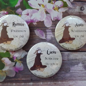 Peut inclure: Trois badges ronds de couleur crème avec un design de chapeau de sorcier marron et de hibou. Les badges comportent du texte en français, notamment des noms et des phrases comme "Parrain" et "Sorcier du Jour". Le fond comprend des fleurs et une surface en bois.