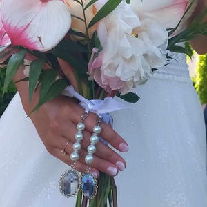 May include: A wedding bouquet with white and pink peonies, anthurium, and foliage. The bouquet is tied with a white ribbon and decorated with two pearl and silver charm bracelets. The bracelets have oval charms with pixelated designs.
