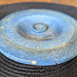 May include: A round, blue ceramic lid with a central knob and visible cracks. The lid has a glossy finish and a slightly uneven surface, suggesting it is handmade. The color is a vibrant blue with hints of brown and yellow.