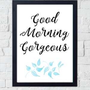 Könnte beinhalten: Gerahmter Kunstdruck mit dem Schriftzug "Good Morning Gorgeous" in schwarzer Schrift. Der Druck zeigt hellblaue Aquarellblätter am unteren Rand. Der Rahmen ist schwarz, vor einer weißen Backsteinmauer.