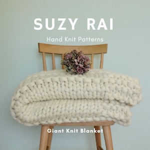 May include: A white chunky knit blanket draped over a wooden chair. The blanket is folded in half and the chair is against a light blue wall. The text "SUZY RAI Hand Knit Patterns Giant Knit Blanket" is below the blanket.