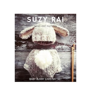 May include: A hand-knitted baby bunny ears hat set in a light brown colour. The set includes a hat with bunny ears and a matching nappy cover with a white pom-pom tail. The set is laid out on a wooden surface with knitting needles.