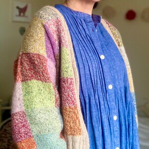Op de afbeelding: Een veelkleurig patchwork gebreid vest met een beige rand. De mouwen hebben vierkanten in rood, groen, roze en oranje. Het vest wordt gedragen over een blauwe jurk met plooien en witte knopen.