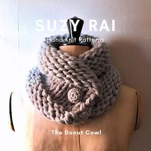 May include: A chunky, gray knit cowl with a button detail, displayed on a mannequin. The text "SUZY RAI Hand Knit Patterns" and "The Donut Cowl" are visible. The cowl is a soft, warm accessory.