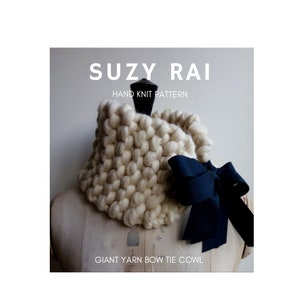 May include: A white knitted cowl with a large black bow tie. The cowl is made with chunky yarn and has a textured look. The text "SUZY RAI HAND KNIT PATTERN GIANT YARN BOW TIE COWL" is printed below the cowl.