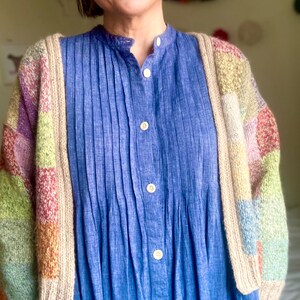 Op de afbeelding: Een blauwe overhemdjurk met verticale plooien wordt gedragen onder een kleurrijk vest in patchworkstijl. Het vest heeft blokken pastelkleuren, waaronder roze, groen en geel. De jurk heeft een opstaande kraag en witte knopen.