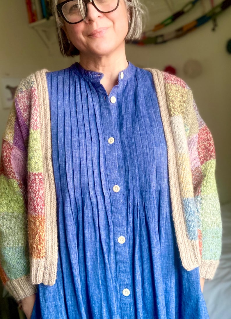 Op de afbeelding: Een blauwe overhemdjurk met verticale plooien wordt gedragen onder een kleurrijk vest in patchworkstijl. Het vest heeft blokken pastelkleuren, waaronder roze, groen en geel. De jurk heeft een opstaande kraag en witte knopen.