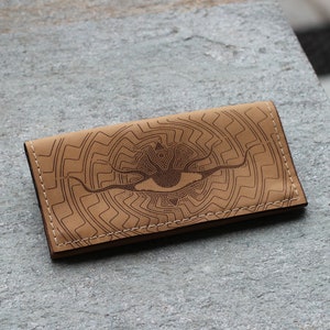 Op de afbeelding: Een bruine leren portemonnee met een gedetailleerd oogontwerp en een geometrisch patroon. De portemonnee heeft een gestikte rand.