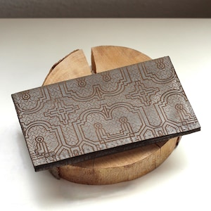 May include: A rectangular brown leather wallet featuring a dark brown embossed geometric pattern with repeating arched and cross-shaped motifs. The wallet has visible stitching along the edges and a flat, slim profile with a textured surface finish.