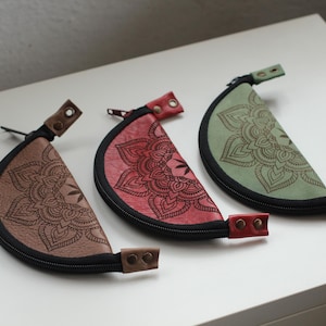 Pode incluir: Três bolsas de couro em forma de meia-lua em marrom, vermelho e verde. Cada bolsa apresenta um zíper preto e um design decorativo de mandala. As bolsas têm uma alça de couro em cada extremidade.