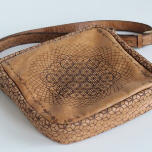 May include: A brown leather crossbody bag with a geometric design and a flower of life pattern. The bag has a long adjustable strap.